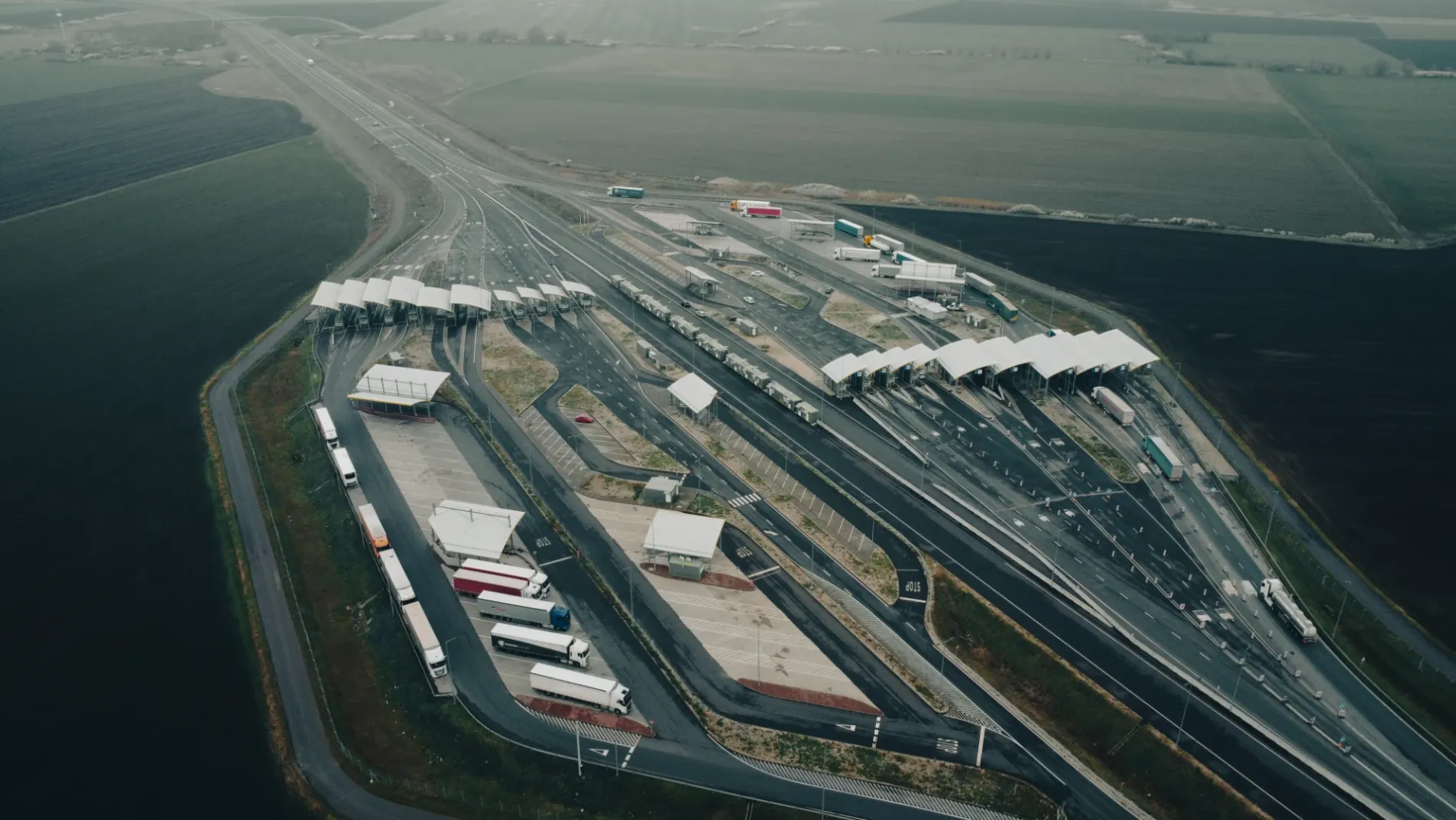 Aerial drone view of large Hungary border crossing complex with multiple truck and car lanes, customs booths and surrounding agricultural fields
